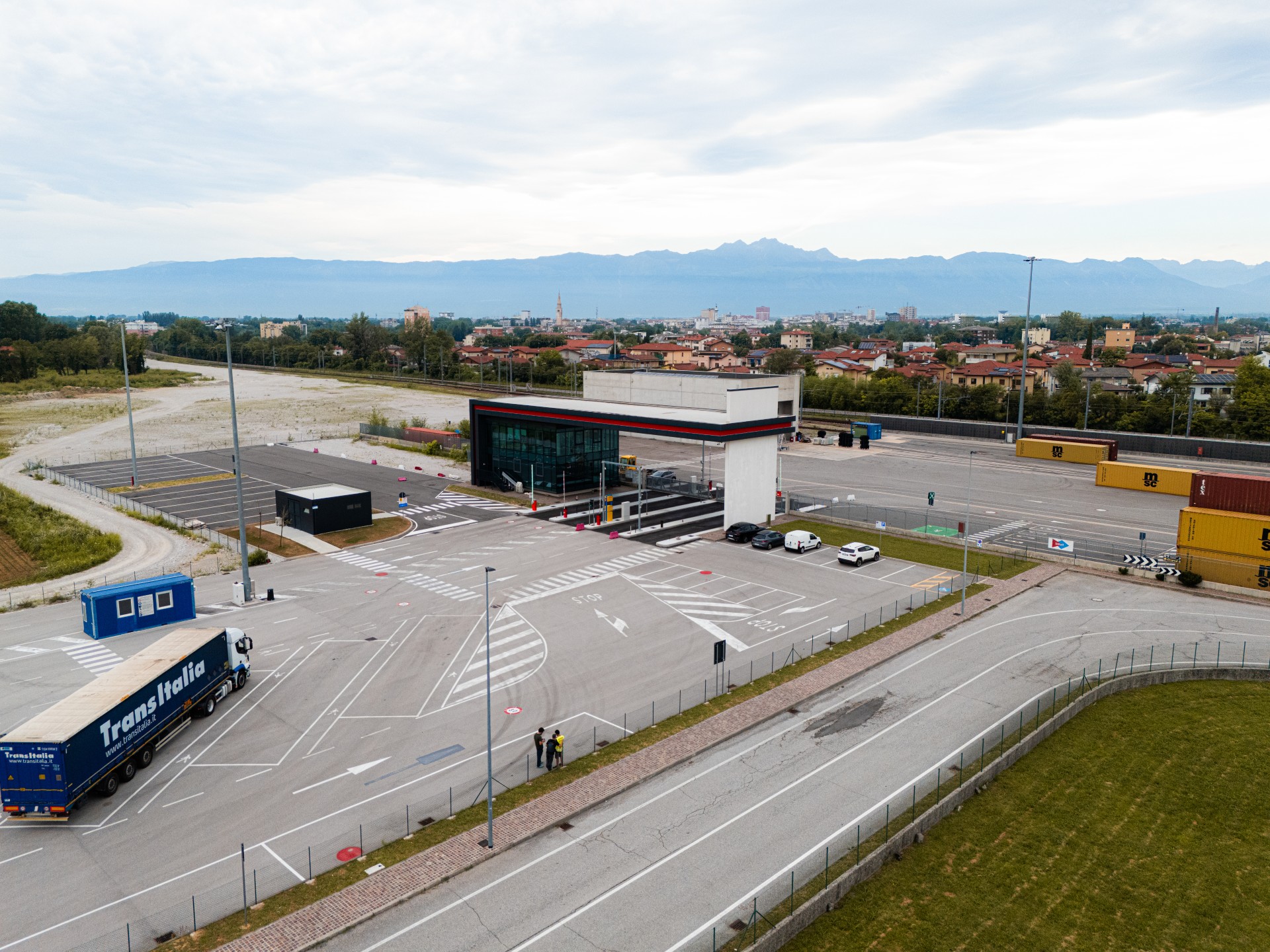 Veduta aerea del Parcheggio Europa: l'ingresso con la barriera di accesso e un autoarticolato sul piazzale di sosta, con i container del terminal e le montagne sullo sfondo